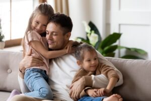 Pleased dad sit on couch with kids, after long separation missed his two little 7s daughters, closing his eyes, hugs them enjoying moment. Family bond and love, Happy Father Day celebration concept