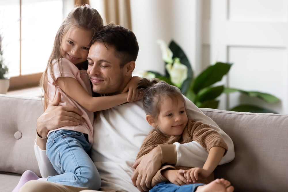 Pleased dad sit on couch with kids, after long separation missed his two little 7s daughters, closing his eyes, hugs them enjoying moment. Family bond and love, Happy Father Day celebration concept